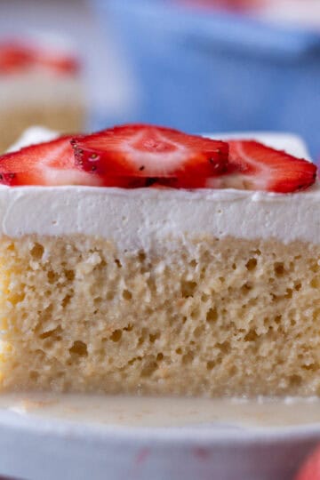 A plate with a slice of tres leches cake, showing the fluffy sponge inside, topped with whipped cream and sliced strawberries.