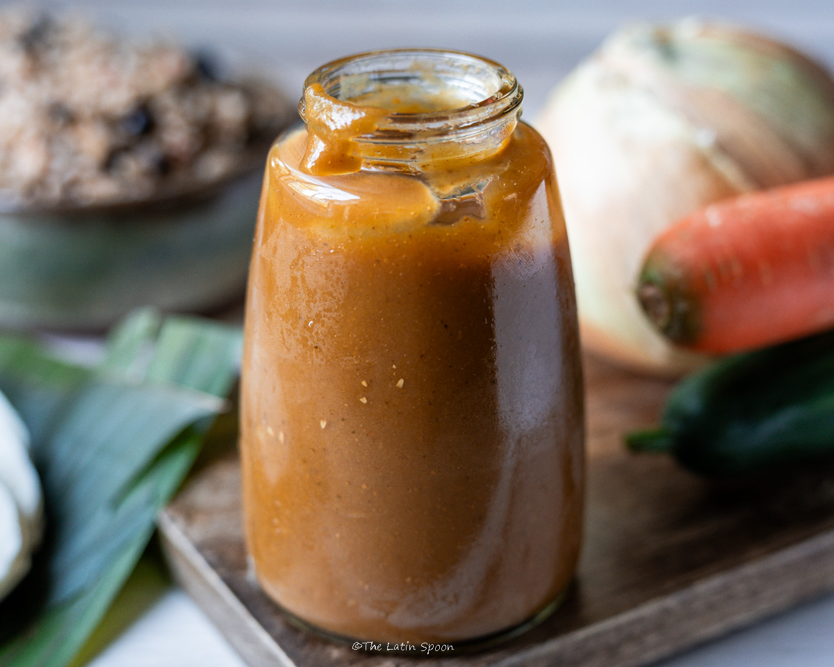 A glass bottle of homemade Lizano sauce on a wooden board, next to an onion, a carrot, and a bell pepper, with a plate of gallo pinto in the background.