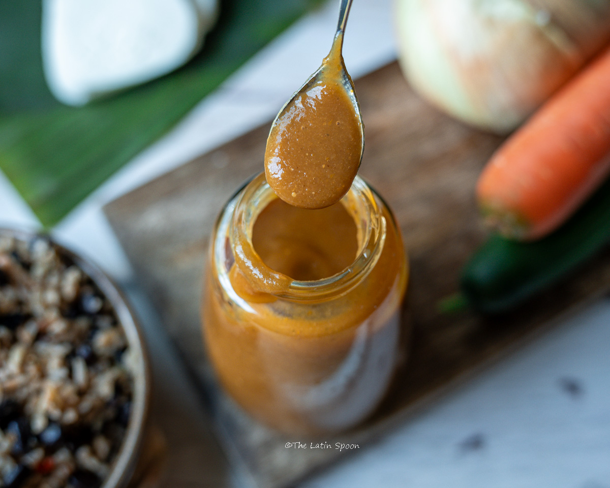 A glass bottle filled with homemade Lizano-style sauce on a wooden board, next to an onion, a carrot, and a bell pepper, with a plate of gallo pinto in the background. A spoon is also shown displaying the sauce’s texture and consistency.