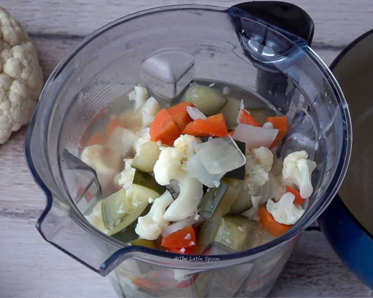 A blender pitcher with the diced vegetables, next to a cauliflower and a pot.