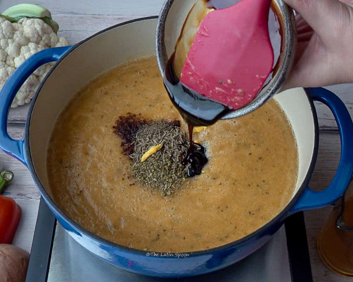 A pot with the blended vegetable mixture and the seasonings on top, while a spatula adds molasses from a small cup.
