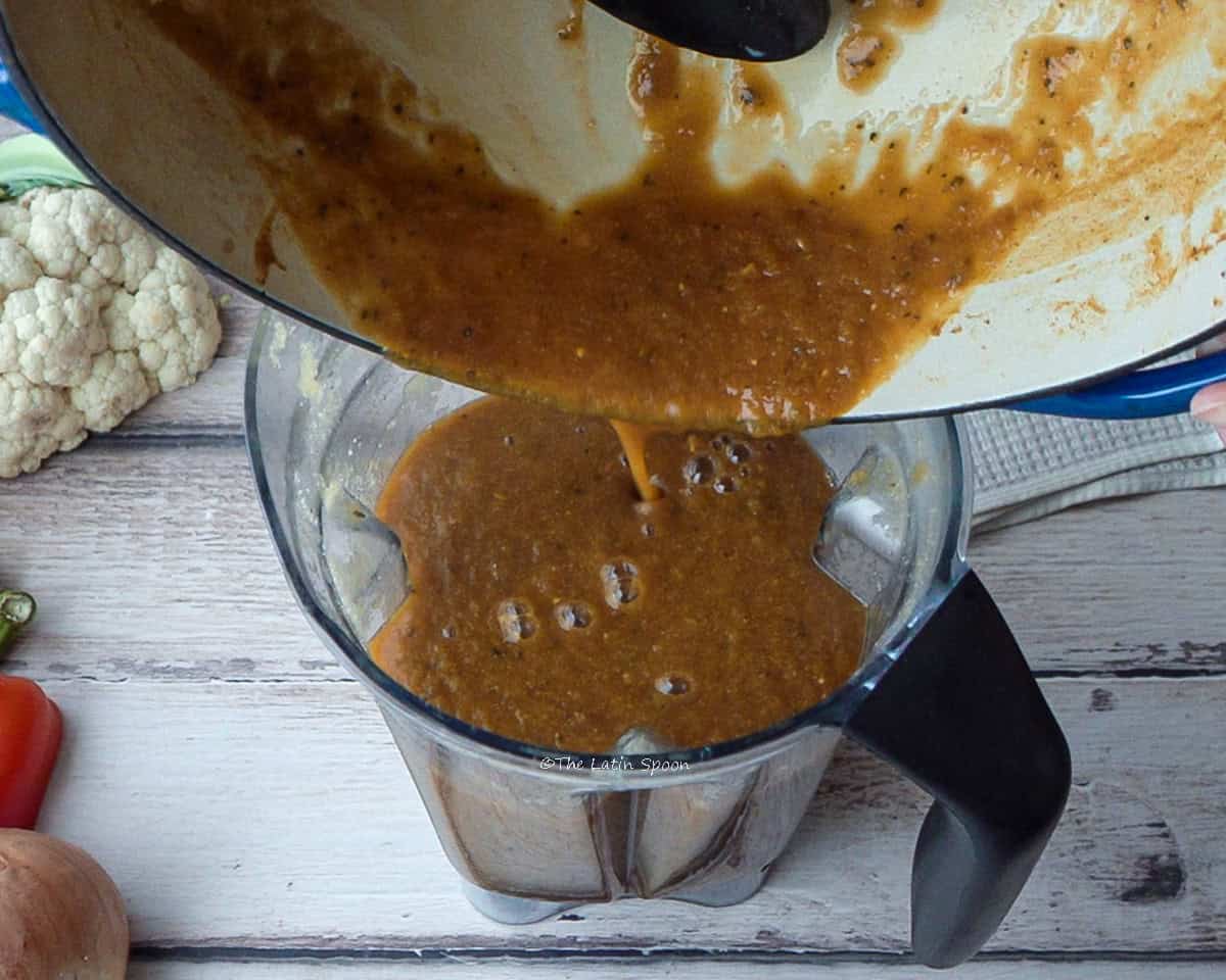 A pot pours the mixture into the blender jar; outside, a cauliflower, a bell pepper, and an onion can be seen.