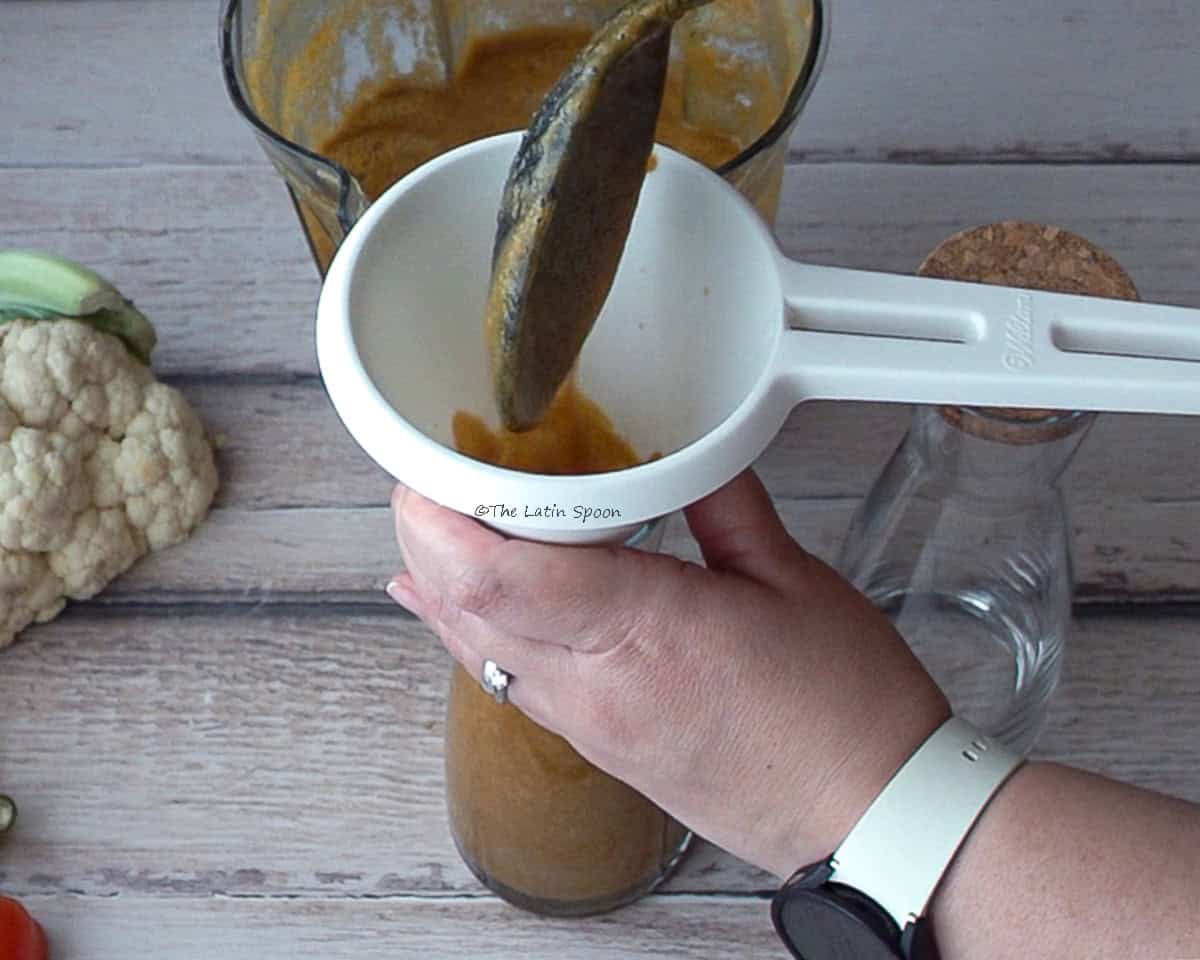 One hand holds a container with a funnel on top, while the other pours the sauce using a spoon. In the background, there is a blender pitcher, a cauliflower, and an empty container beside it.