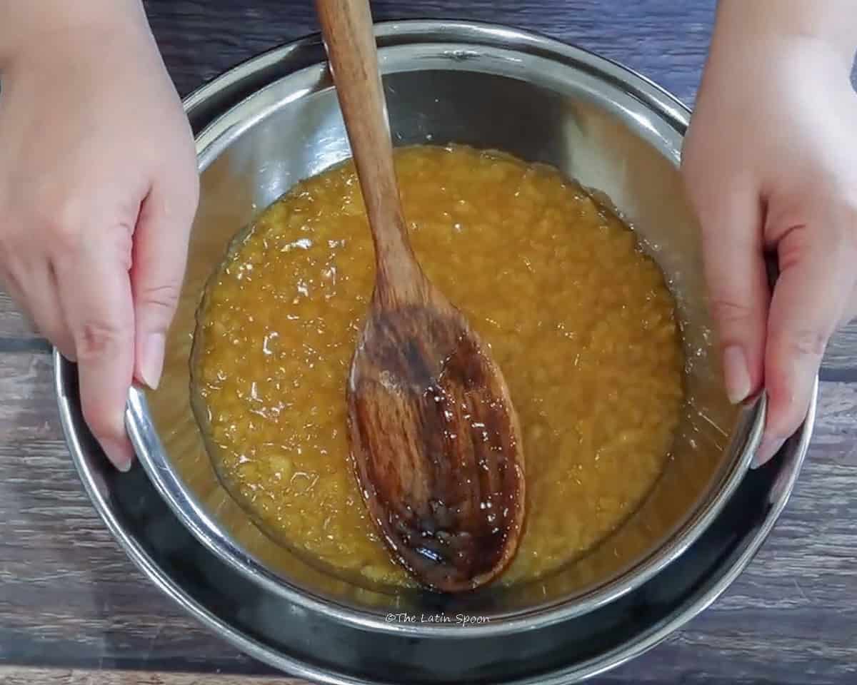 A pot with the finished pineapple jam is shown resting in a container filled with ice, along with a wooden spoon.