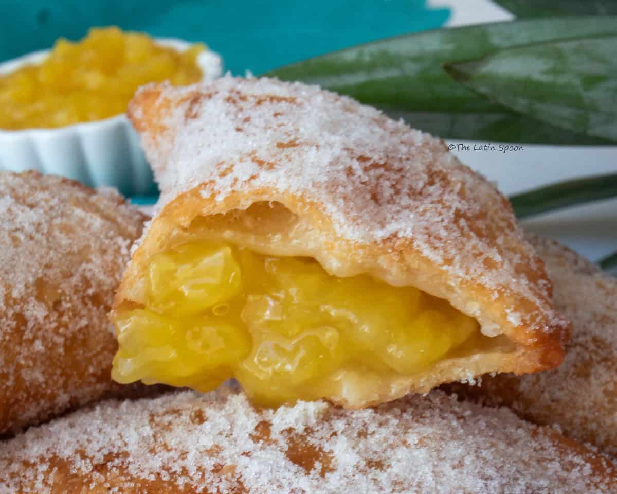 Photograph of a stack of sugar-glazed empanadas, with one empanada on top cut in half, showing the pineapple filling overflowing.