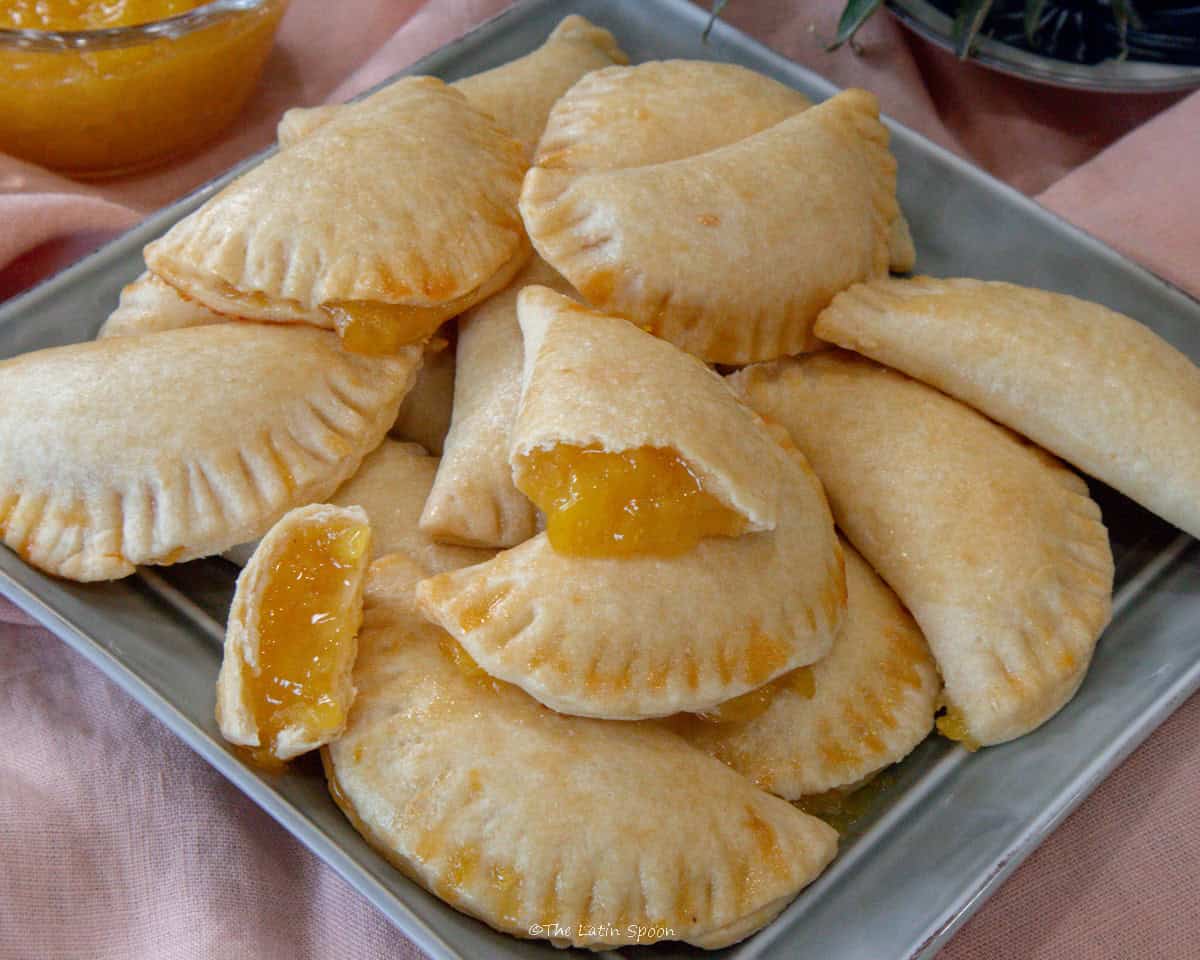 A tray of pineapple empanadas is shown on a tablecloth. In the front, one empanada is cut in half, showing its pineapple filling, and in the background, the crown of a pineapple can be seen as part of the decoration.