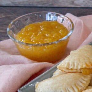 A small glass cup containing pineapple jam is shown, with round-shaped empanadas in the background of the photograph.