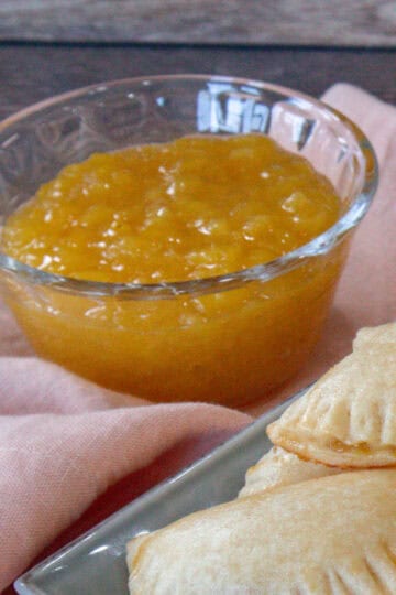 A small glass cup containing pineapple jam is shown, with round-shaped empanadas in the background of the photograph.