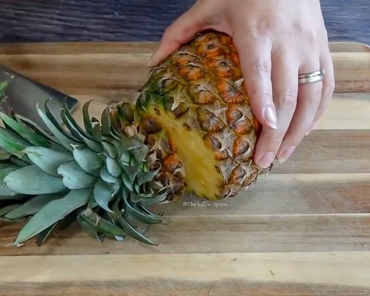 Photograph showing a wooden cutting board where the crown of a pineapple is being cut off.