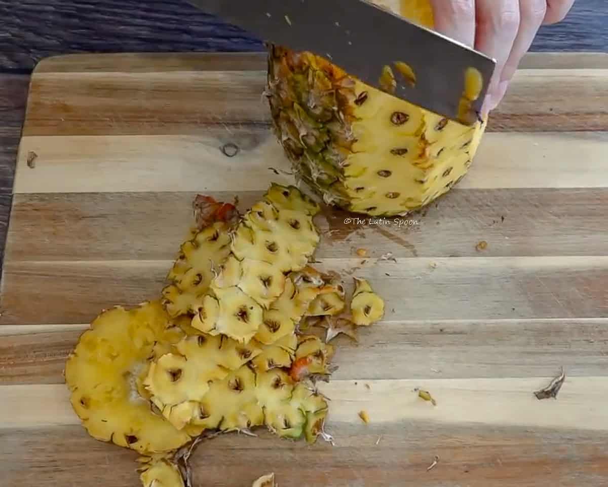 Photograph of a pineapple on a wooden cutting board, being carefully peeled with a knife, while leaving the eyes on the pineapple.