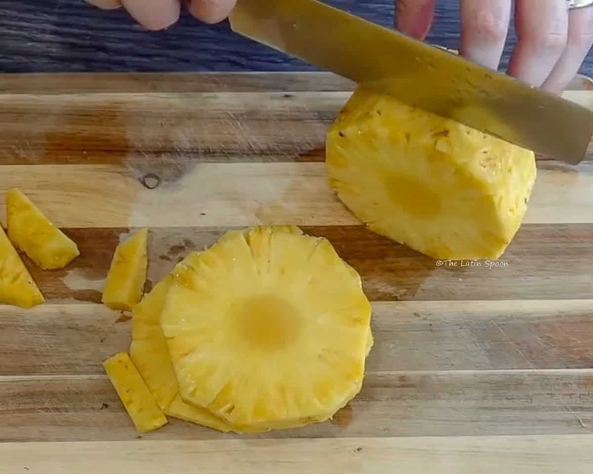 Photograph of a wooden cutting board, some pineapple slices, and a knife cutting the pineapple.
