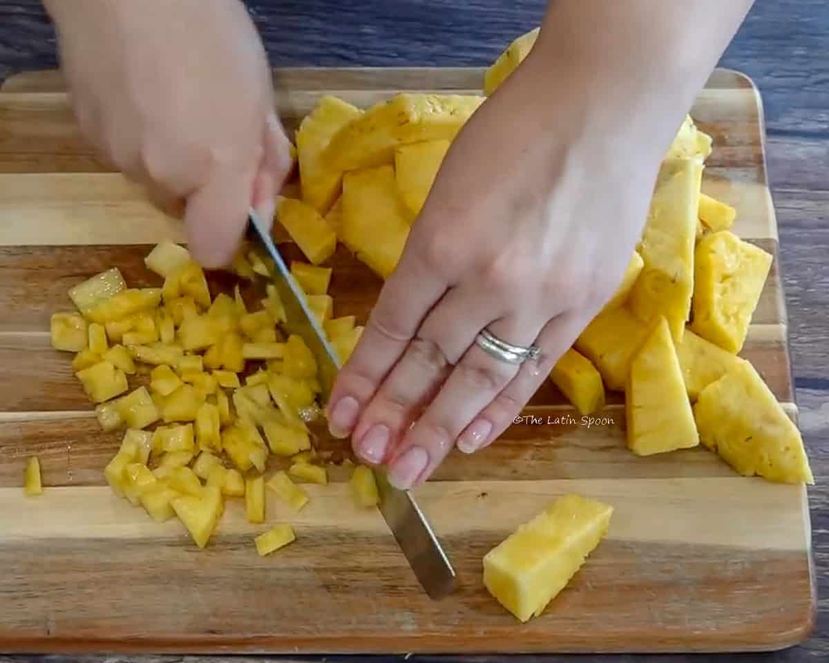 Photograph showing a wooden cutting board with pineapple cut into small cubes, and hands holding a knife while dicing it.