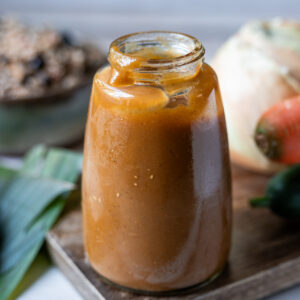 Una botella de vidrio con salsa Lizano casera sobre A wooden cutting board next to an onion, a carrot, and a bell pepper, with a plate of gallo pinto in the background.