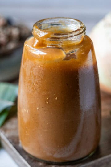 Una botella de vidrio con salsa Lizano casera sobre A wooden cutting board next to an onion, a carrot, and a bell pepper, with a plate of gallo pinto in the background.