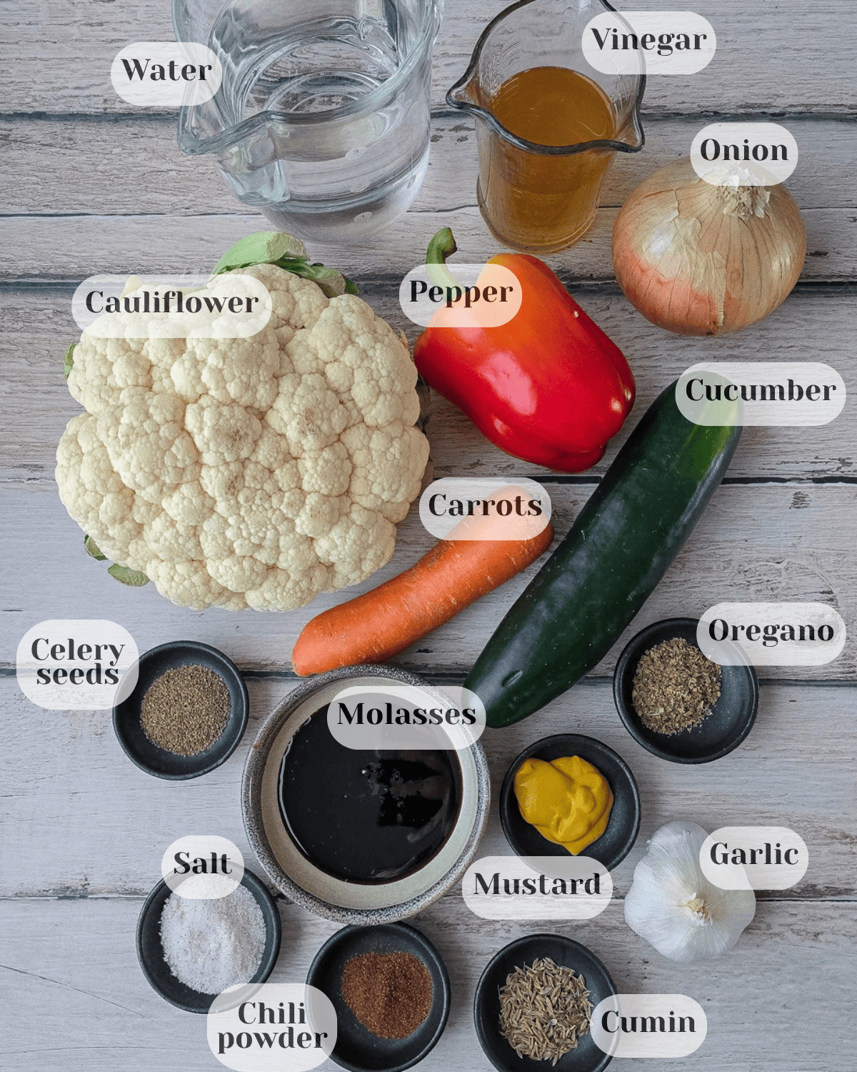The ingredients viewed from above: a cup of water, a small pitcher of vinegar, an onion, a bell pepper, cauliflower, a carrot, a cucumber, black pepper, molasses, oregano, mustard, garlic, salt, hot pepper, and cumin.