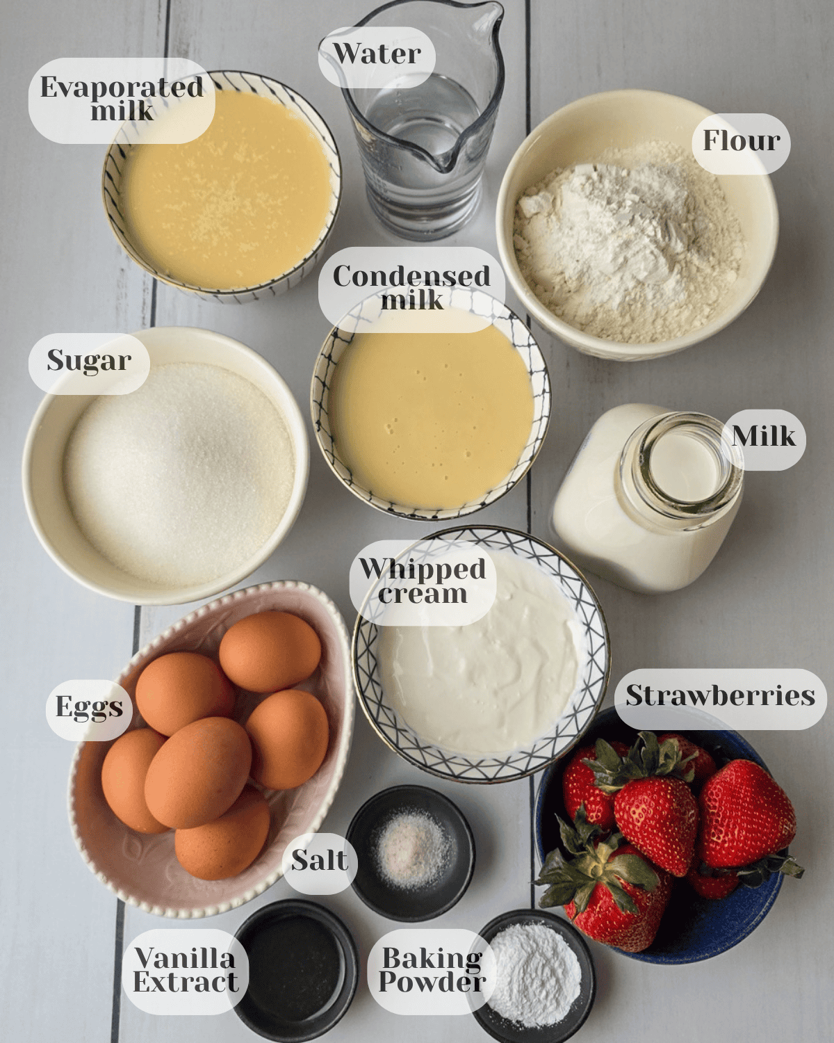 All the ingredients needed for the recipe: a small jar of water, a small bowl of evaporated milk, a small bowl of sweetened condensed milk, a small bowl of flour, a small bowl of sugar, a small pitcher of regular milk, a small bowl of heavy cream, a plate with six eggs, strawberries, salt, baking powder, and vanilla extract served in small bowls.