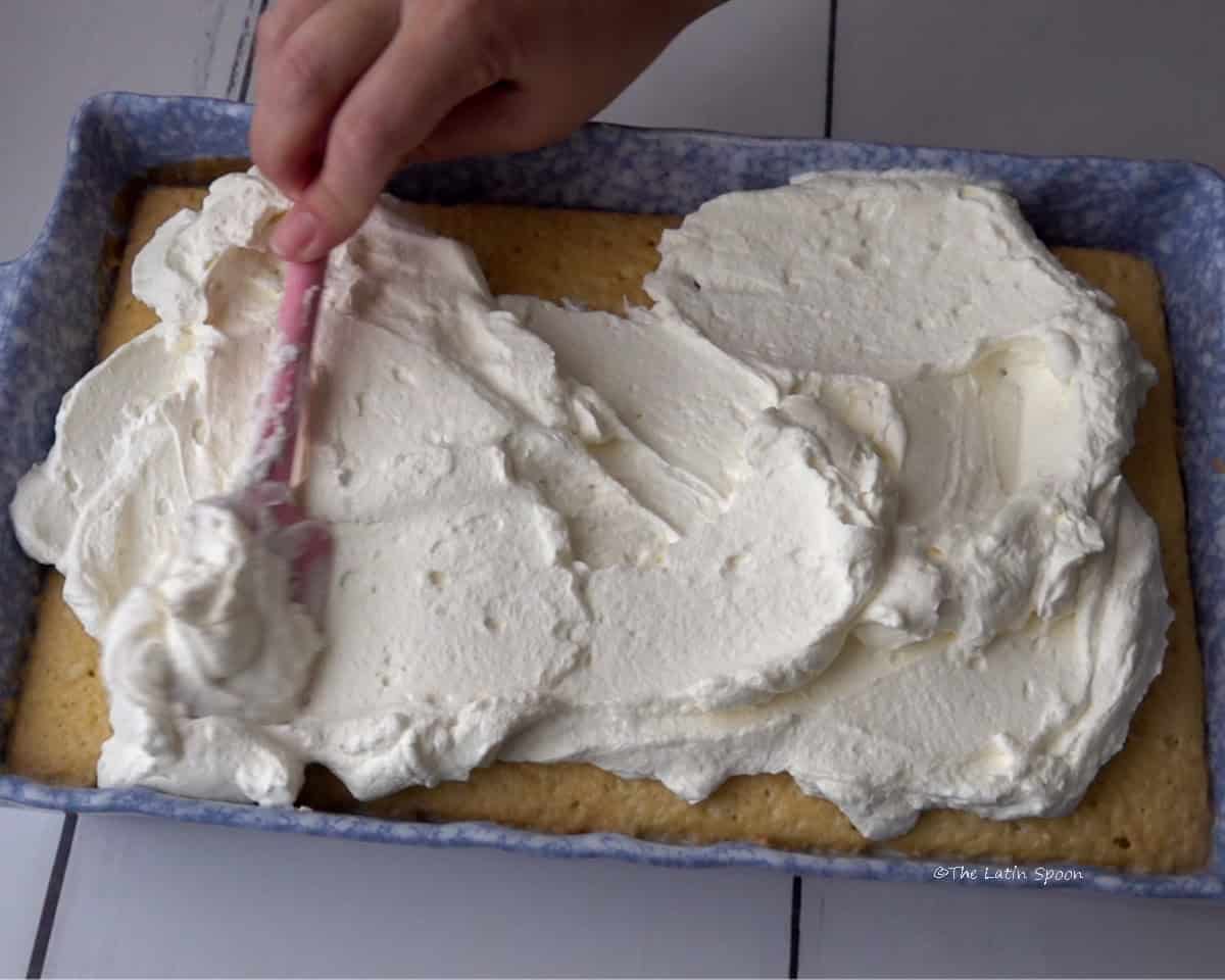 A baking dish with the cake inside, while a hand uses a spatula to spread whipped cream on top.
