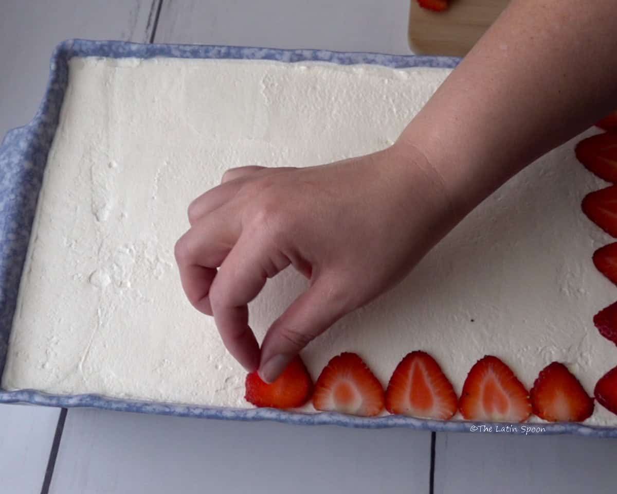 A baking dish containing the cake decorated with whipped cream, while a hand places strawberry slices on top.