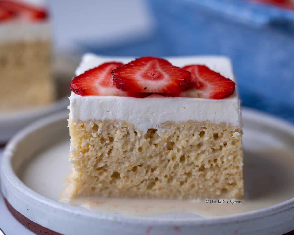 A plate with a slice of tres leches cake, showing the fluffy sponge inside, topped with whipped cream and sliced strawberries.