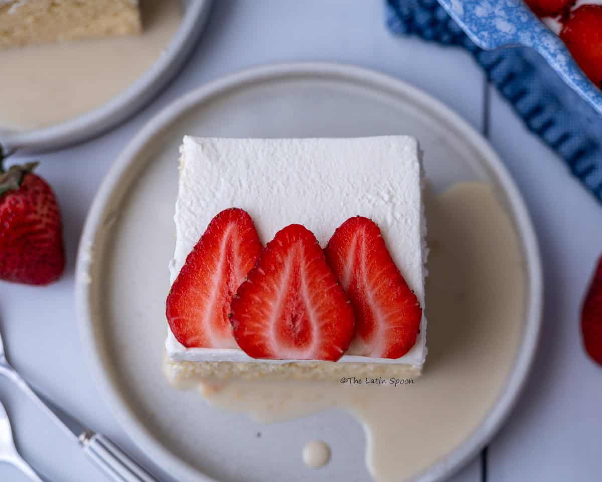 A top-down view of a plate with a slice of tres leches cake topped with whipped cream and three strawberry slices. Some of the milk mixture is visible at the bottom of the plate. In the background, there is another plate served the same way, with a whole strawberry placed beside it.