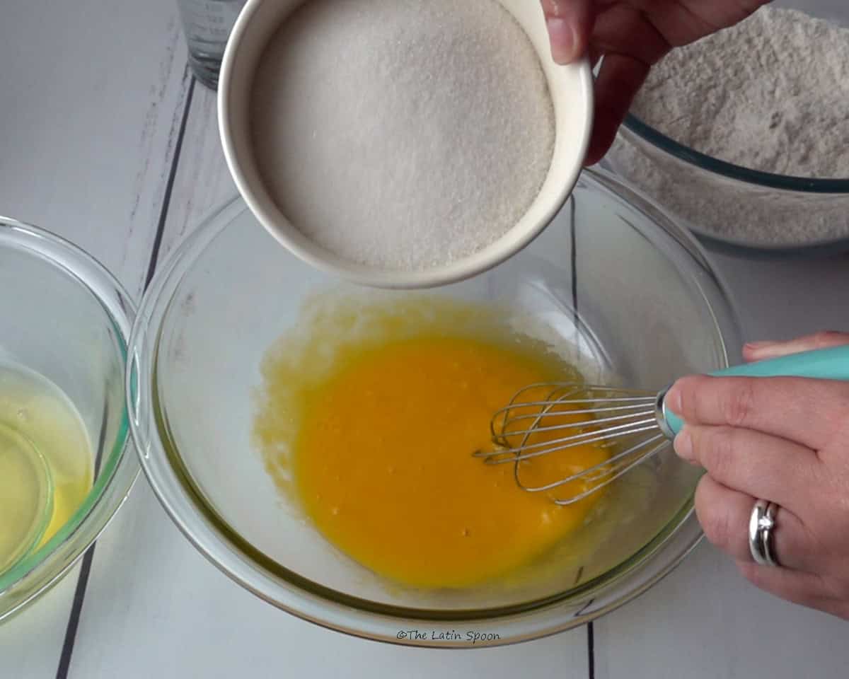 A bowl containing the egg yolks, with the right hand holding a whisk. The left hand holds a small bowl of flour while pouring it into the mixture. Beside it, there is a small bowl with the egg whites, and another bowl of flour can be seen in the background.