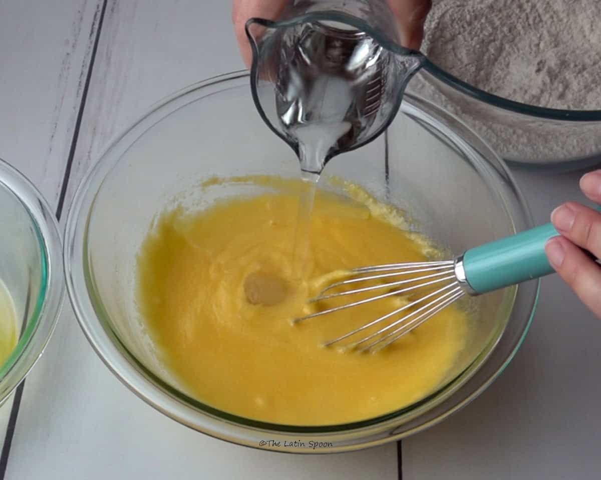 A bowl containing the egg yolk and sugar mixture. The left hand holds a whisk, while the right hand pours water from a small pitcher into the bowl. A bowl of flour can be seen in the background.