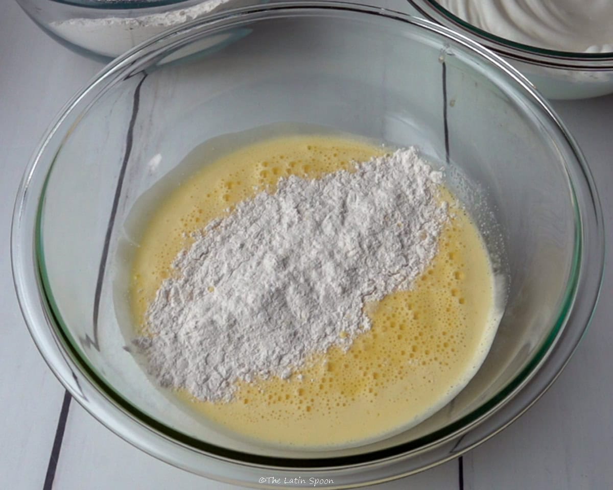 A bowl with the egg yolk, sugar, and water mixture, with some of the flour mixture being added on top.