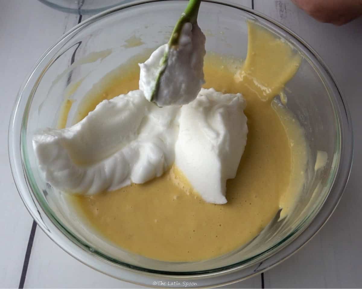 A bowl containing the flour and egg mixture, while a spatula adds fluffy whipped egg whites on top.