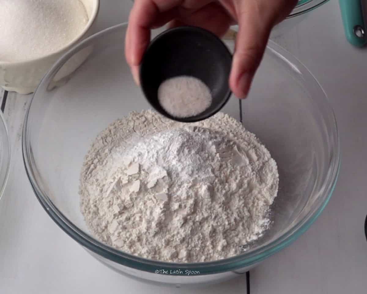 A bowl with flour while a hand holds a small bowl pouring salt into it. In the background, a cup of sugar can be seen.