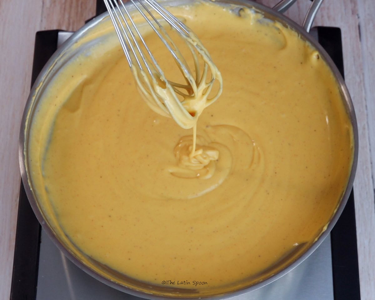 A skillet with finished cheese sauce, while a whisk above it lifts a strand of cheese.