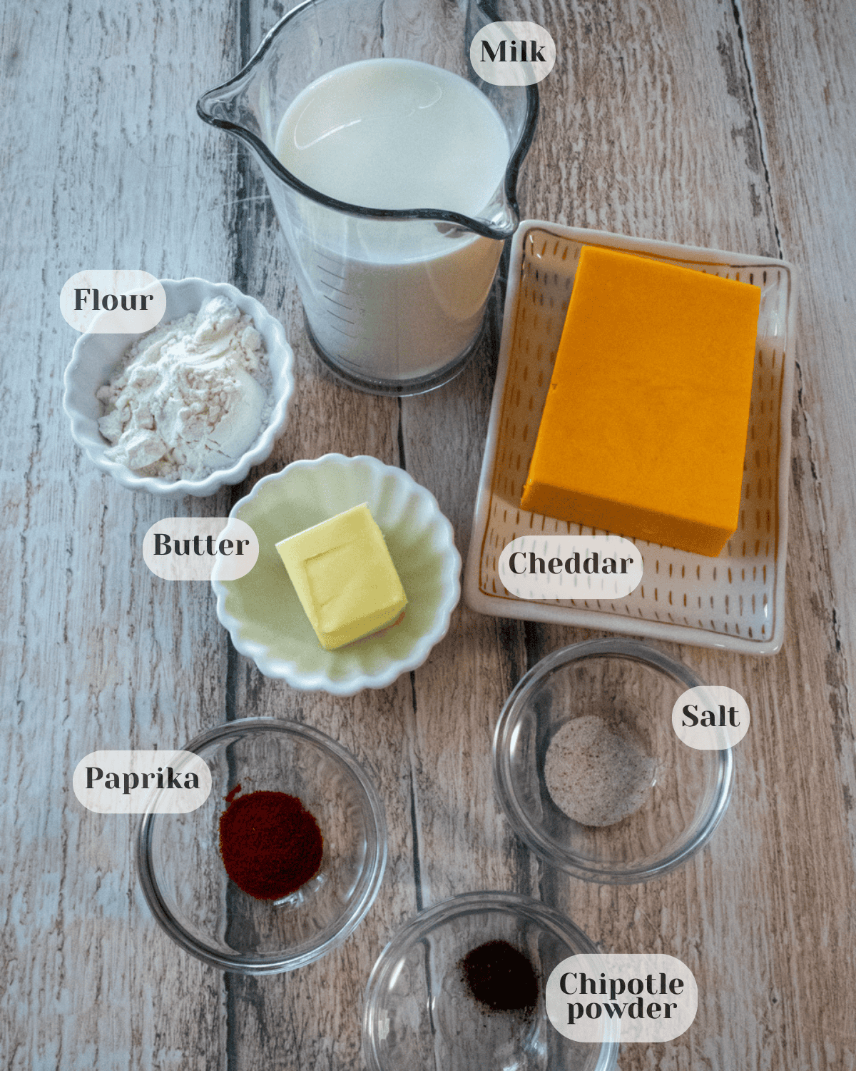 Ingredients grouped together: a small pitcher of milk, a small cup of flour, a quarter stick of butter, a plate with a block of cheddar cheese, and small cups of paprika, chipotle powder and salt.