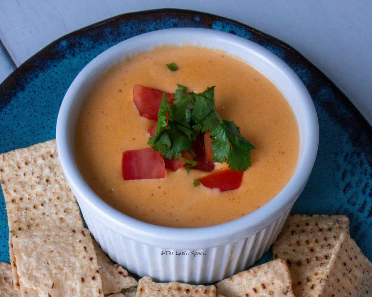 A small cup of cheddar cheese sauce topped with pieces of tomato and cilantro sits on a plate of tortilla chips.