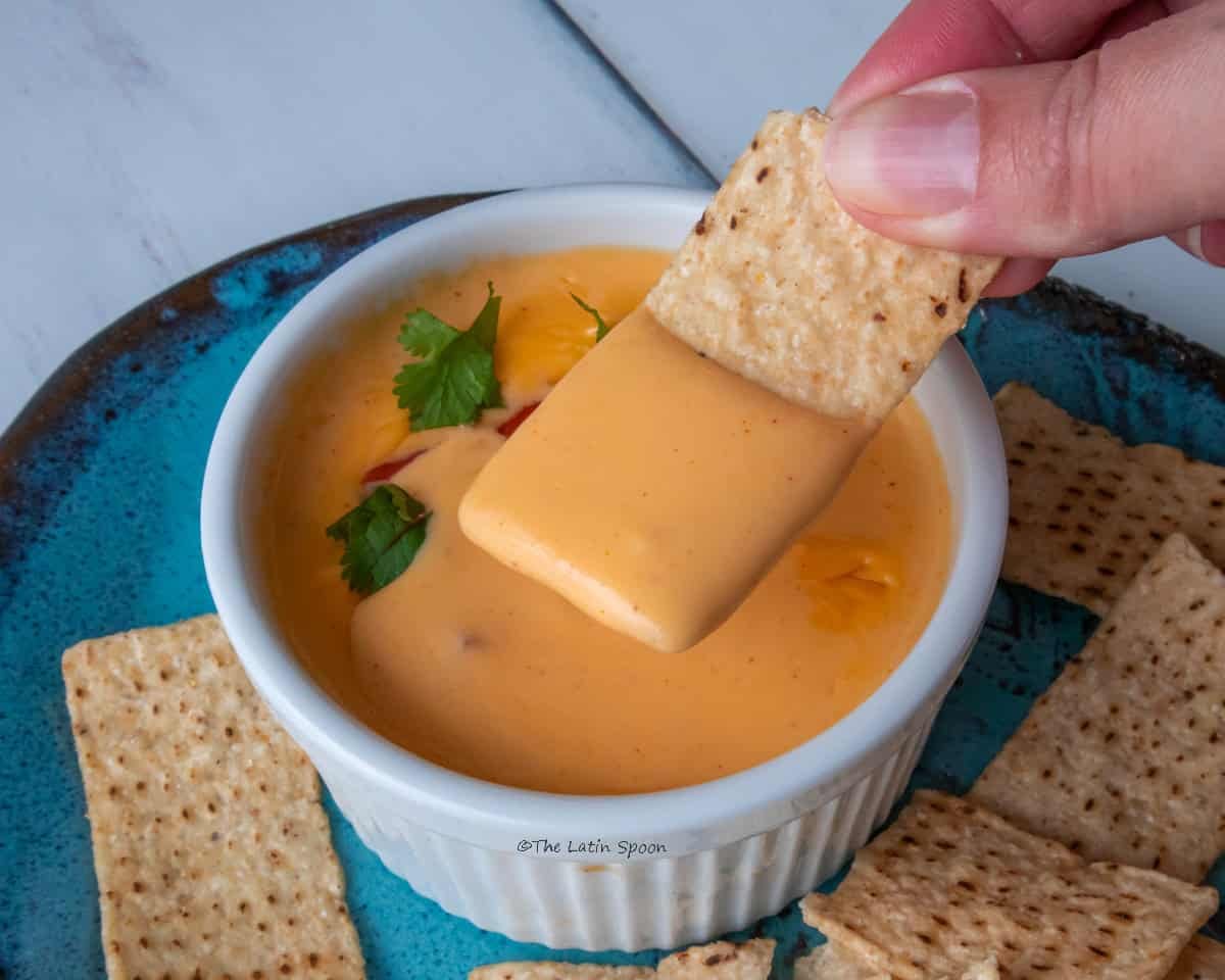 A small cup of cheddar cheese sauce topped with pieces of tomato and cilantro sits on a plate of tortilla chips. A hand holds a freshly dipped tortilla chip above the sauce.
