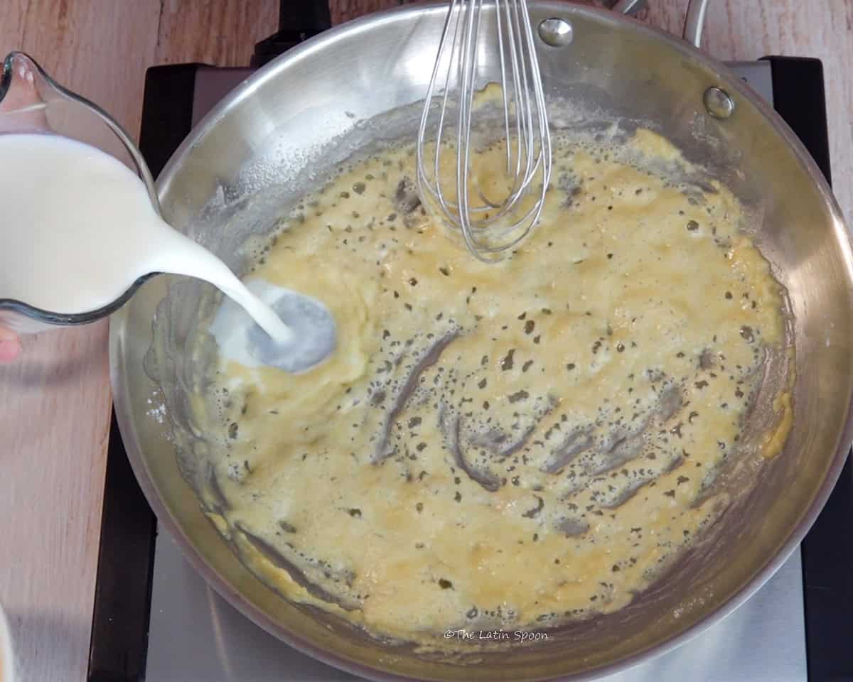 Milk is being poured from a pitcher into a skillet containing a mixture of butter and flour.