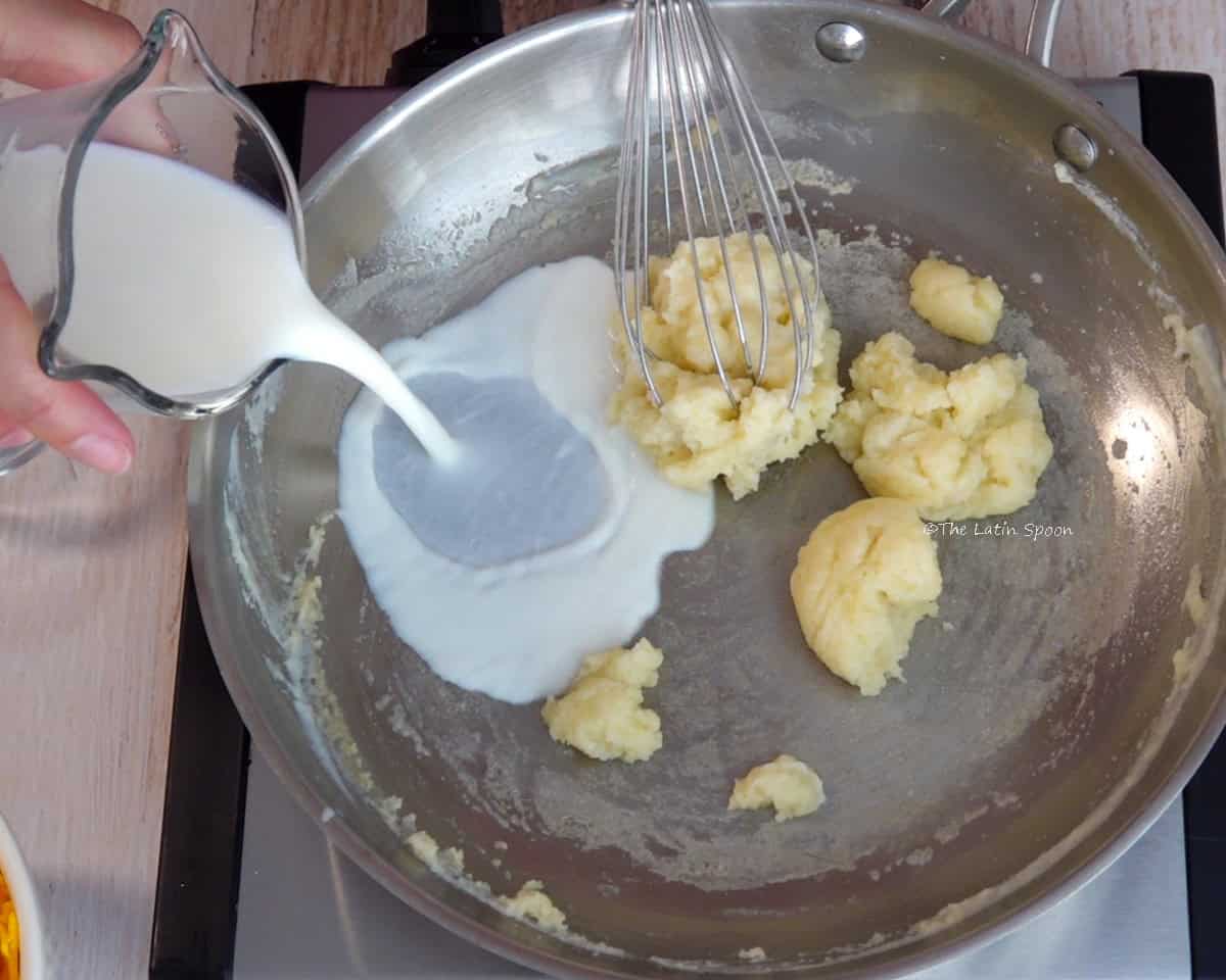 More milk is poured from a pitcher into a skillet containing a thick butter and flour mixture, with some of the mixture clumped around the whisk.