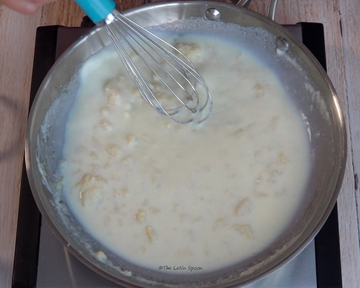 A mixture of milk, butter, and flour with some lumps is being stirred with a whisk in a skillet.