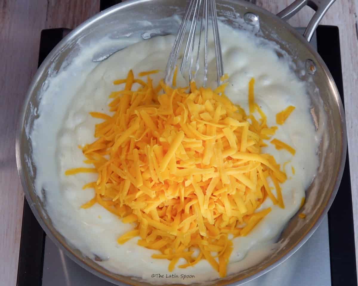 Shredded cheddar cheese is being added to the thick mixture in the skillet.