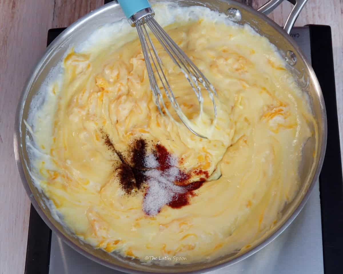 Paprika, salt, and cayenne pepper are being added to a nearly combined mixture of cheese, milk, butter, and flour.
