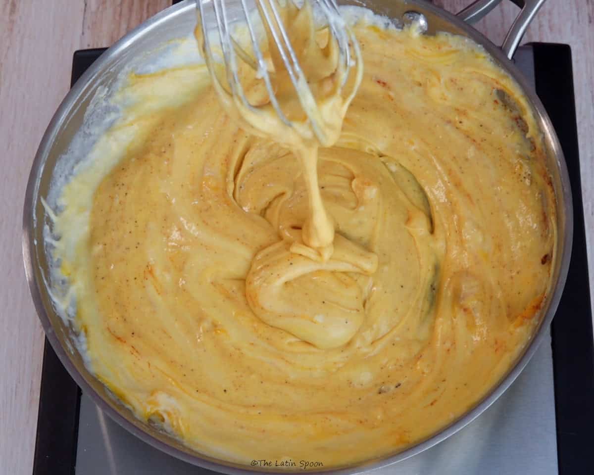 Cheese sauce in a skillet being stirred with a whisk to mix in the seasonings.