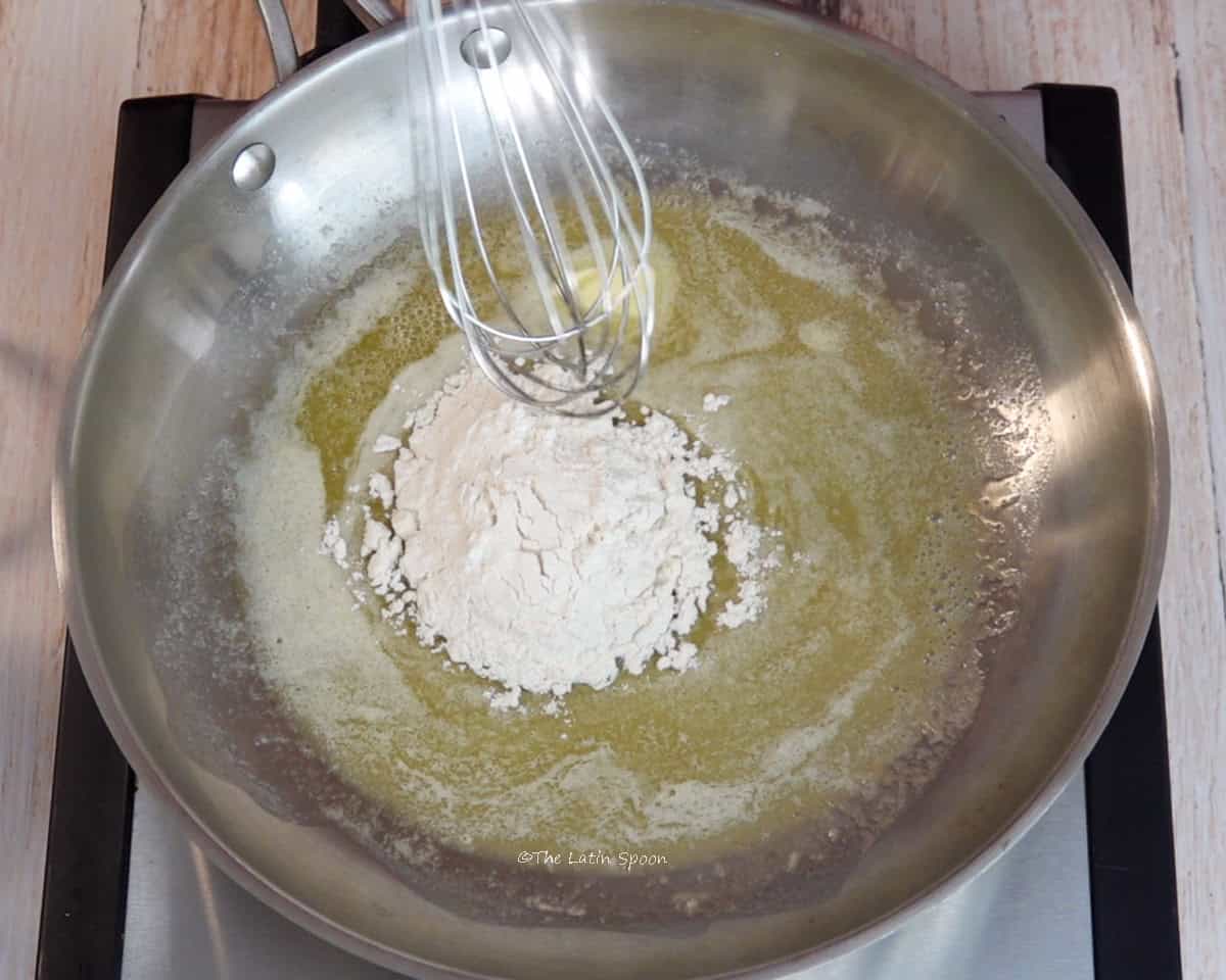 A skillet on a stovetop contains melted butter with some flour in the center and a whisk above it.