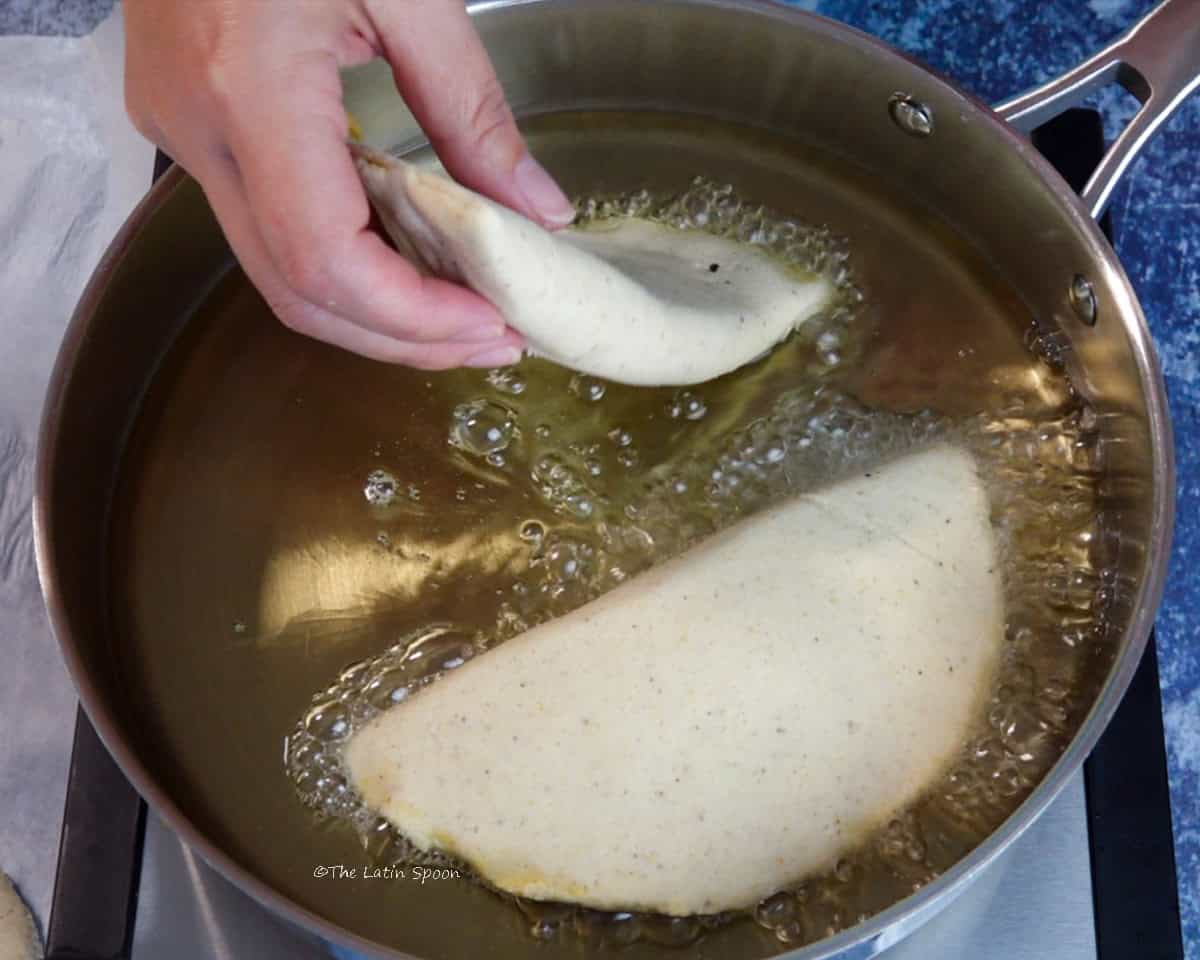 A skillet with hot oil and one empanada frying while a hand places another empanada into the oil.