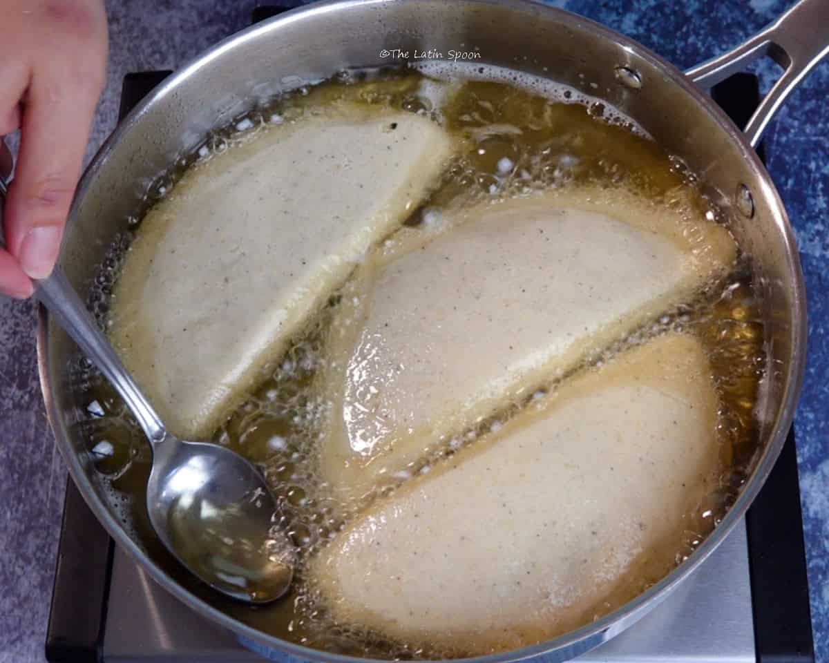 A skillet with three empanadas frying while a hand uses a spoon to pour hot oil over them.