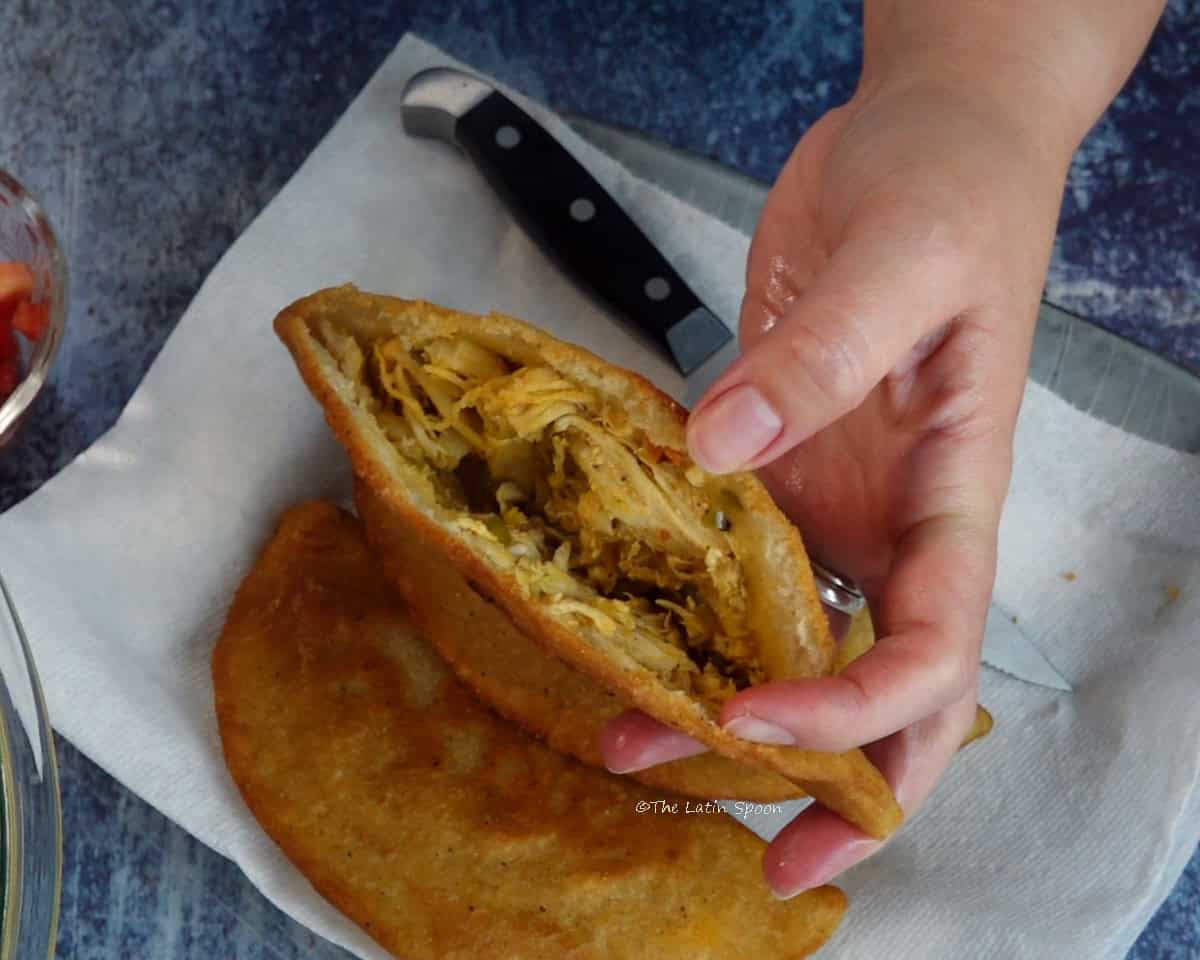 A hand holding an open empanada, with a plate containing two cooked empanadas and a knife in the background.