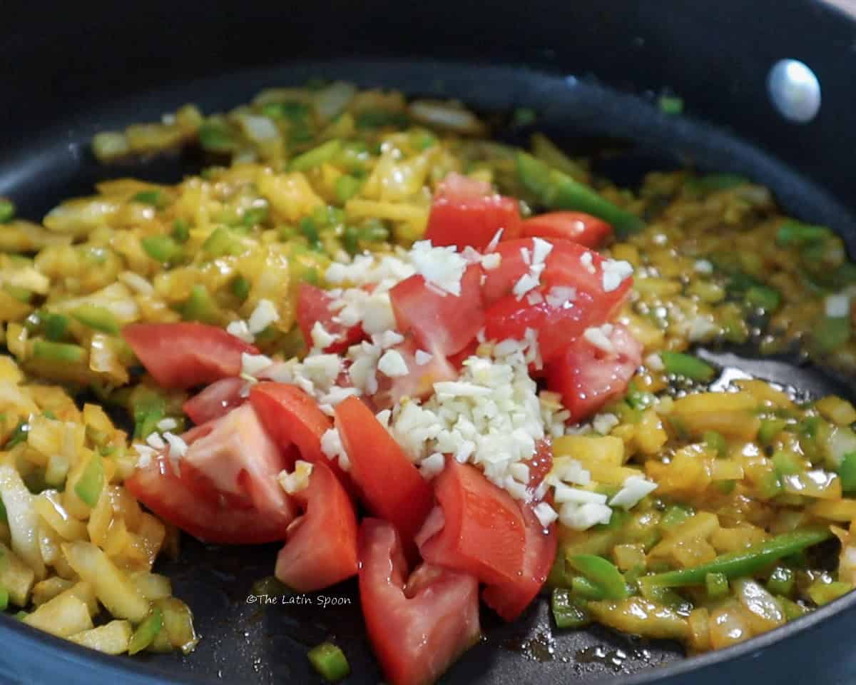 A skillet with a mixture of chopped onion, bell pepper, celery, and other aromatics, topped with pieces of tomato and minced garlic.