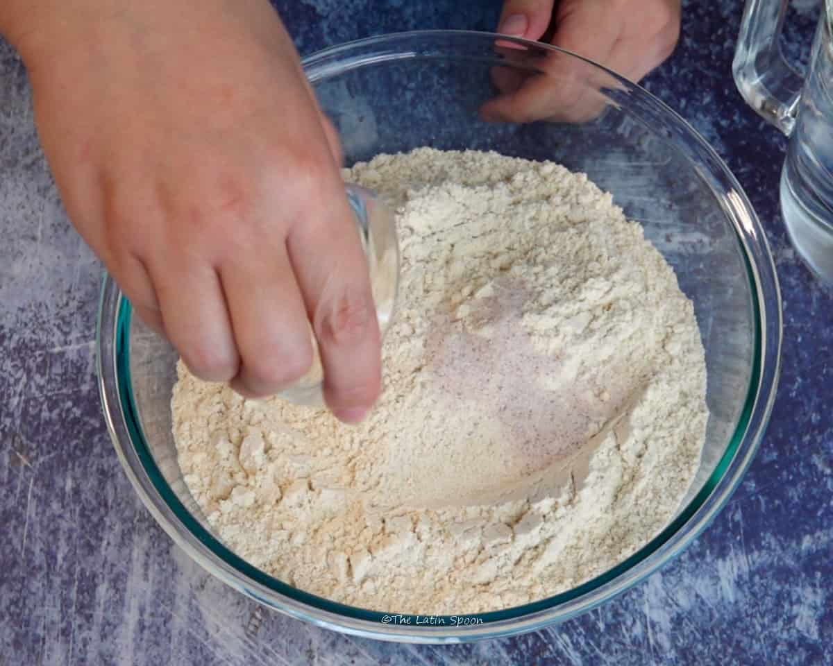 A bowl of dry masa mixture while a hand sprinkles salt over it.