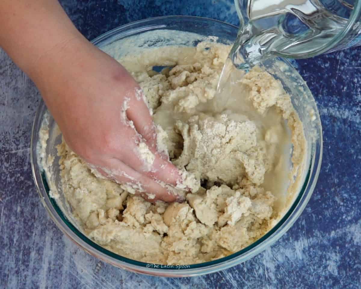 A bowl of masa being mixed by one hand while the other hand pours water from a pitcher.