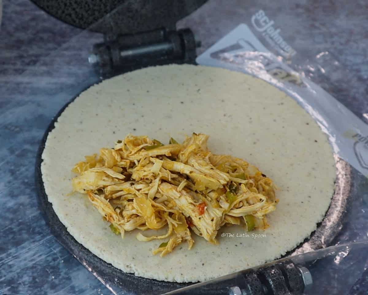 A flattened masa circle filled with shredded chicken on a plastic sheet in a tortilla press, ready to be folded into an empanada.