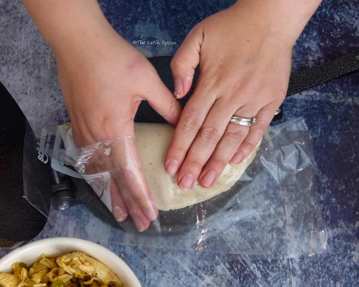 Two hands press one side of the masa over the filling to form the empanada between the plastic sheets.