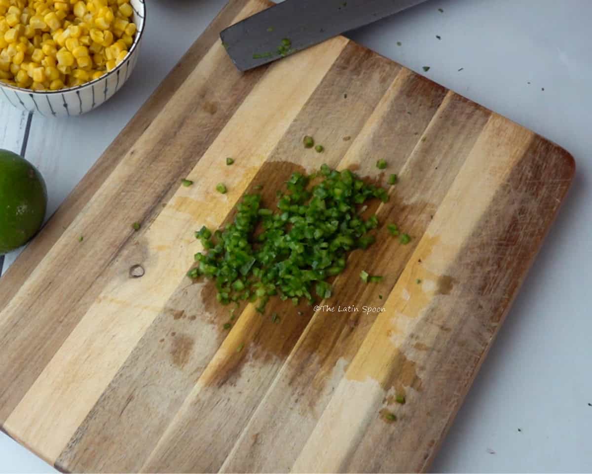 A cutting board with a knife on the side. On the board, there is a jalapeño diced into small cubes, and next to it there is a cup of sweet corn and a lemon.