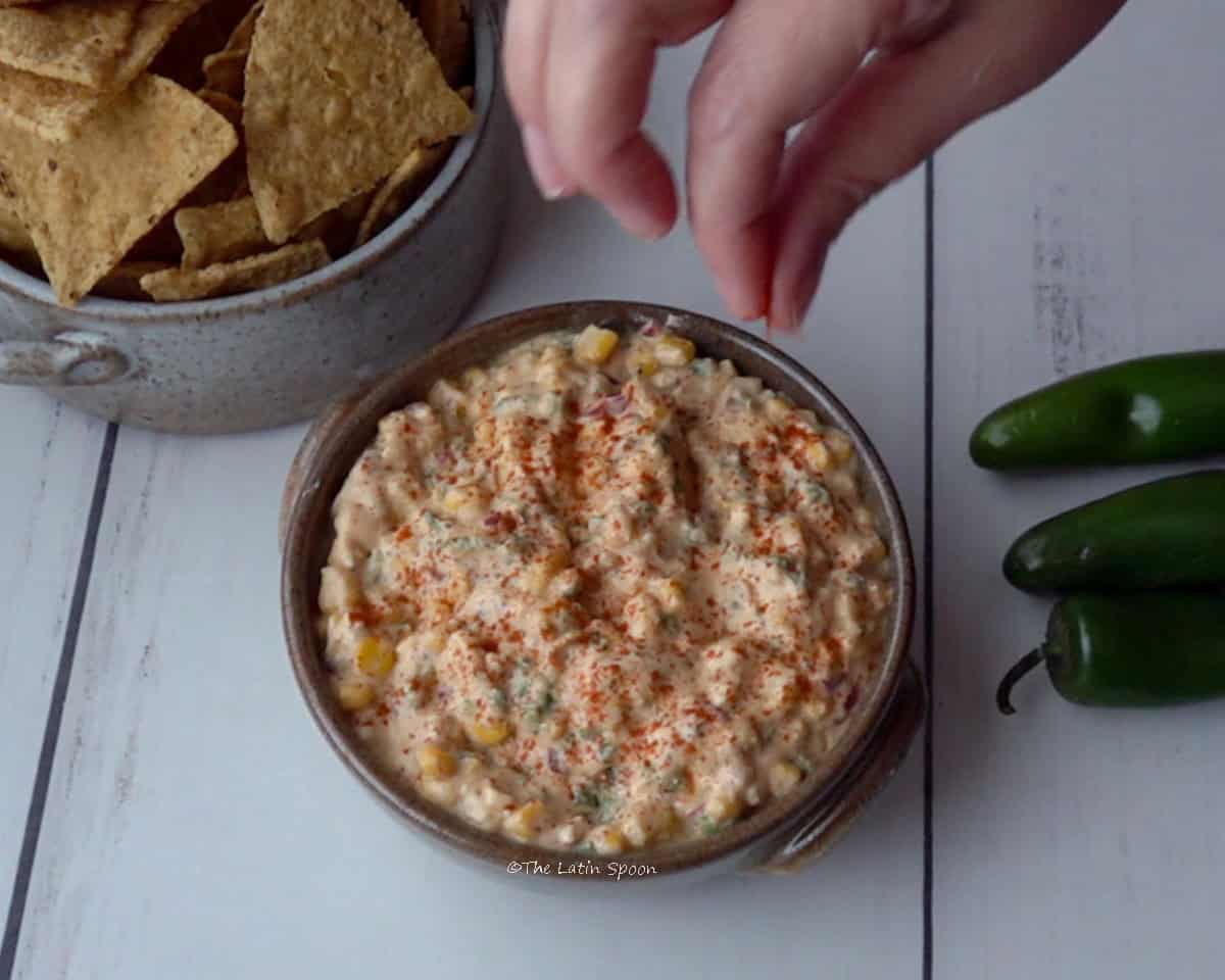 A bowl of corn dip with a hand on top adding paprika to add color. Jalapeños and tortilla chips are shown in the background